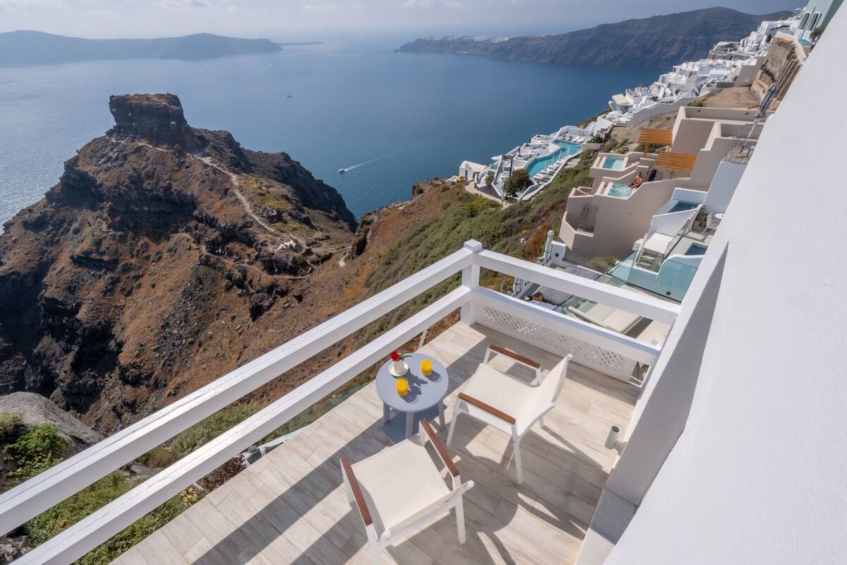 Un balcón con mesa y sillas que da a un paisaje costero con edificios blancos y un risco rocoso.