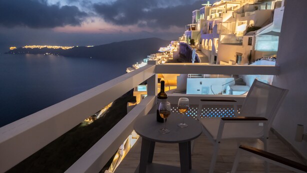 Un balcon avec une table, deux verres et une bouteille, donnant sur une ville côtière au crépuscule.