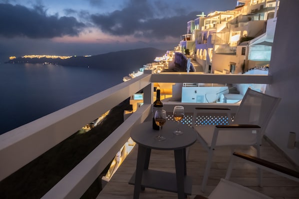 A balcony with a table, two glasses, and a bottle, overlooking a coastal town at dusk. - Anemos Cave Suites (Santorini)
