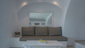 A room with a white sofa, a white coffee table with a fruit bowl, and a small potted plant. - Anemos Cave Suites (Santorini)