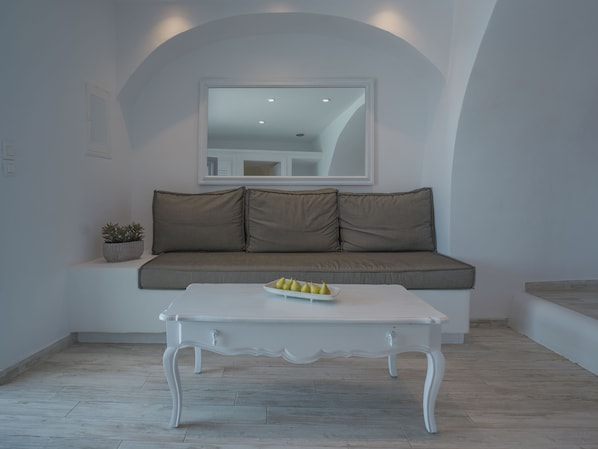 A room with a white sofa, a white coffee table with a fruit bowl, and a small potted plant. - Anemos Cave Suites (Santorini)