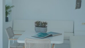 A minimalist room with a white table, two chairs, a small potted plant, and a single window. - Anemos Cave Suites (Santorini)