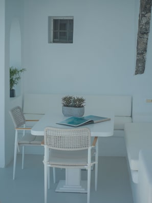 A minimalist room with a white table, two chairs, a small potted plant, and a single window. - Anemos Cave Suites (Santorini)