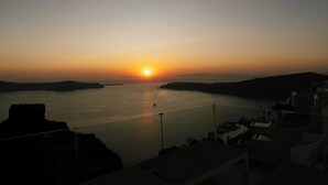 Vista do pôr do sol de uma varanda com vista para um mar calmo e formações terrestres ao longe.