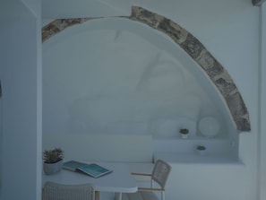 Un interior luminoso con un arco de piedra, una mesa blanca, dos sillas y algunas plantas en macetas.