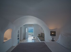 A bedroom with a large bed, a bedside table with a lamp, and a view of a balcony. - Anemos Cave Suites (Santorini)
