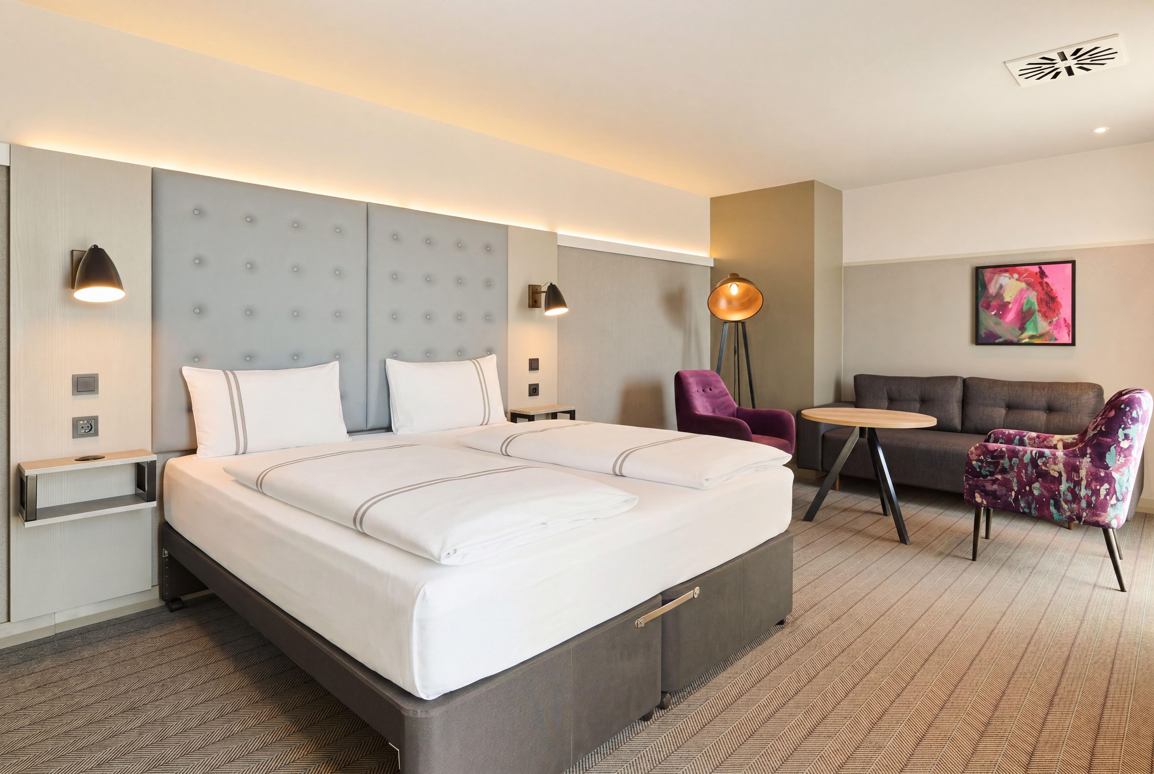 Habitación de hotel con una cama grande, mesitas de noche, una lámpara de pie, una pequeña mesa redonda, un sofá y un cuadro abstracto de colores en la pared.