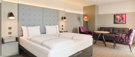 Habitación de hotel con una cama grande, mesitas de noche, una lámpara de pie, una pequeña mesa redonda, un sofá y un cuadro abstracto de colores en la pared.