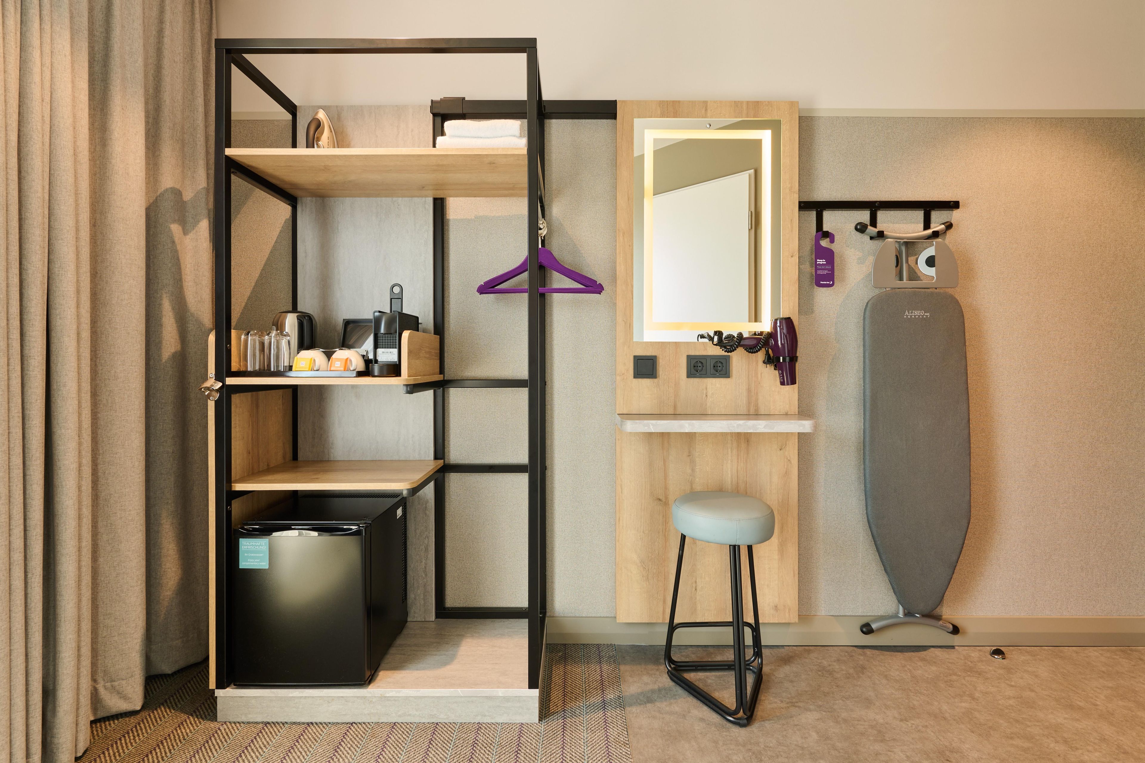 A compact hotel room with a built-in wardrobe, a small seating area, a mirror, and a compact exercise machine.