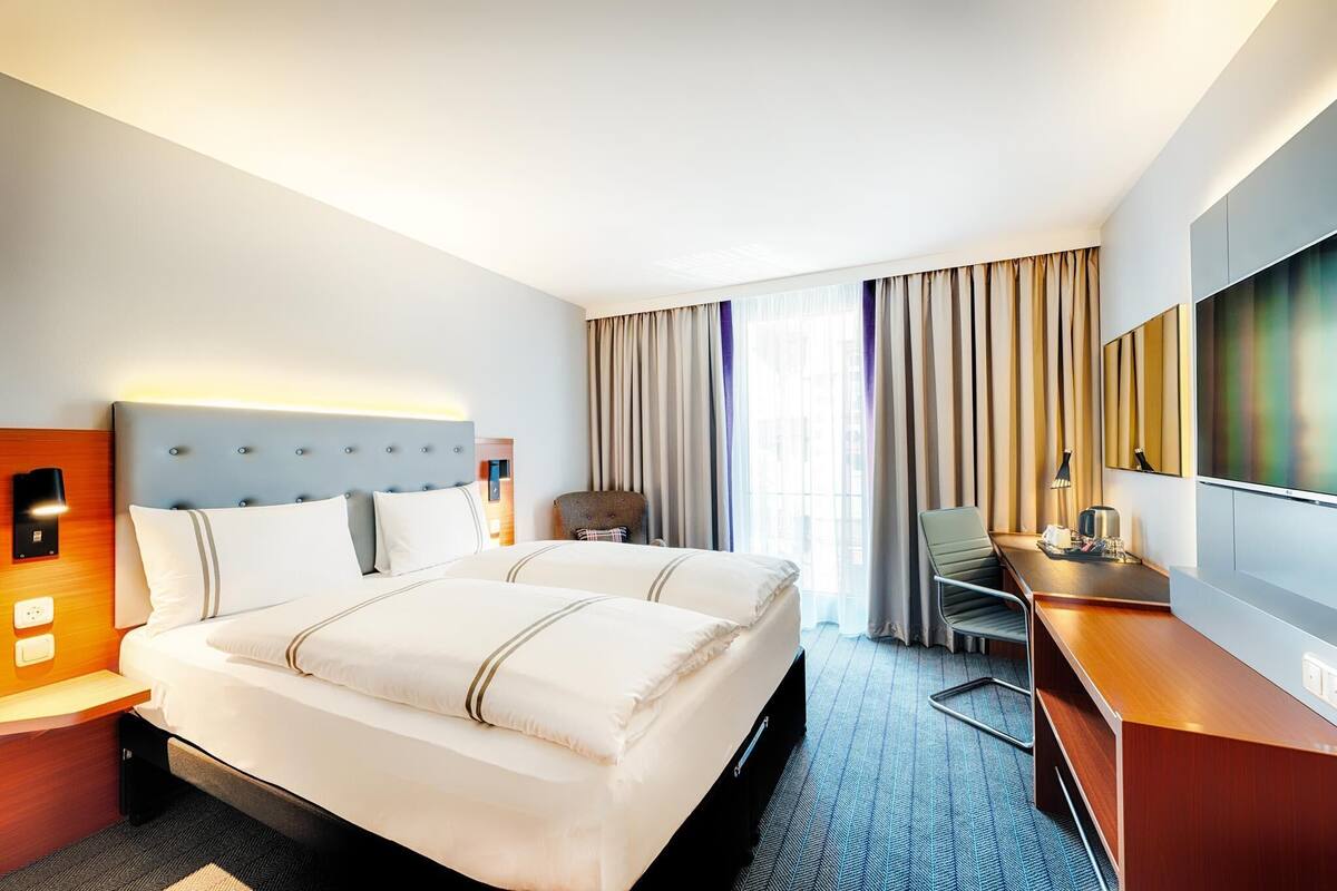 A hotel room with a large bed, a desk, a chair, a TV, and a window with curtains.