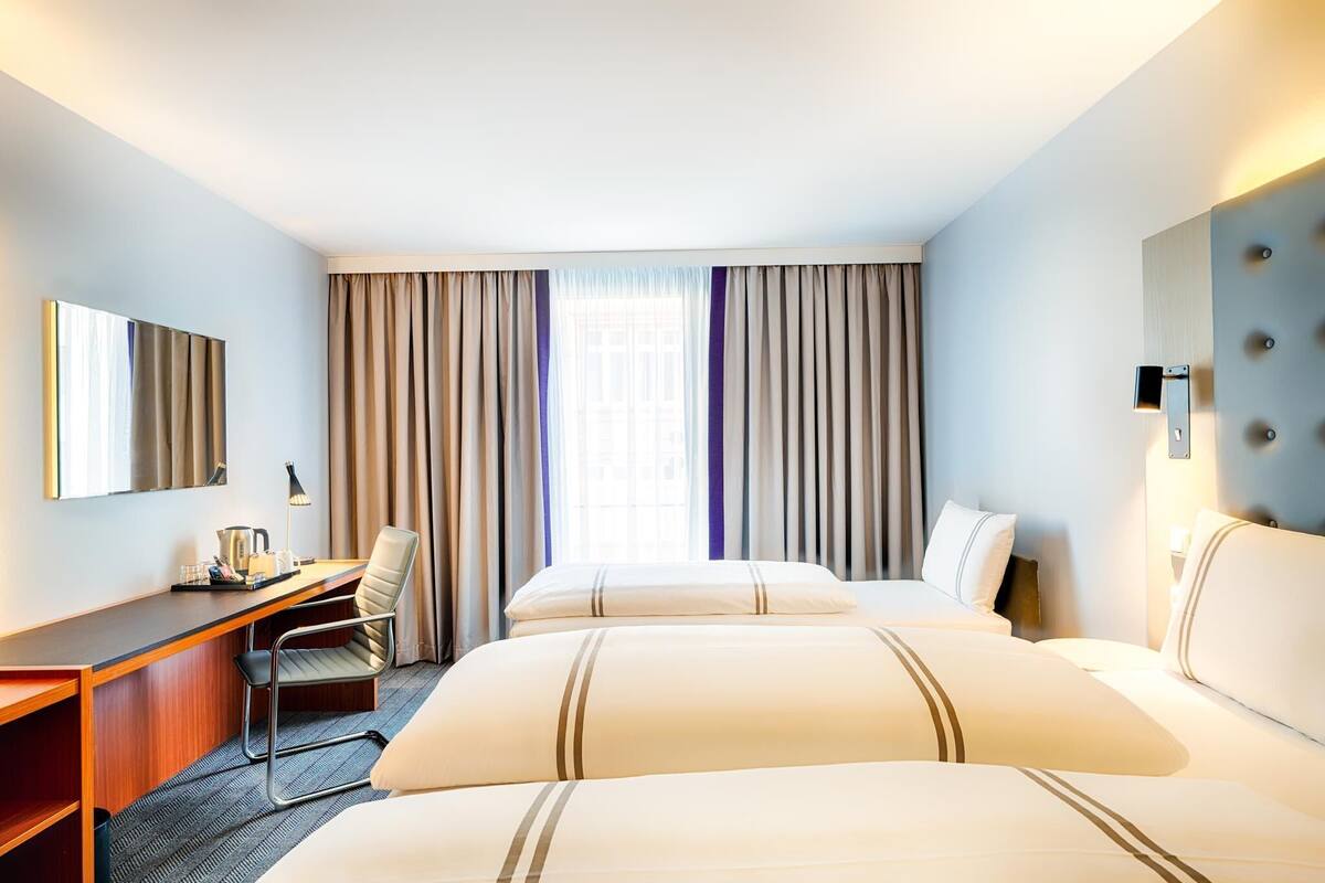 A hotel room with two beds, a desk, a chair, a mirror, and a window with curtains.