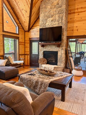 A cozy interior with a stone fireplace, wooden ceiling beams, and a large window. - Stone Mountain Lodge – Spacious 6-Bedroom Forest Cabin (Broken Bow)