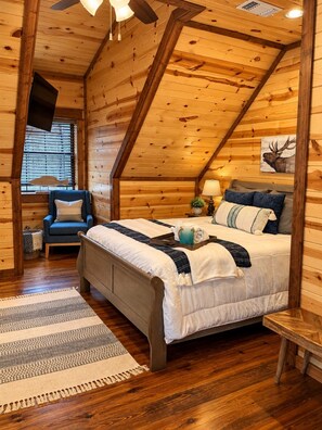 Un dormitorio de cabaña de troncos con cama, una silla, una mesita de noche, una lámpara y un televisor instalado en la pared.