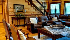 A spacious living room with a wooden staircase, leather furniture, and a large clock on the wall.