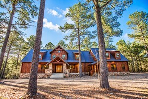 A cabin with a stone foundation, wooden exterior, and a blue metal roof, surrounded by tall pine trees. - Stone Mountain Lodge – Spacious 6-Bedroom Forest Cabin (Broken Bow)
