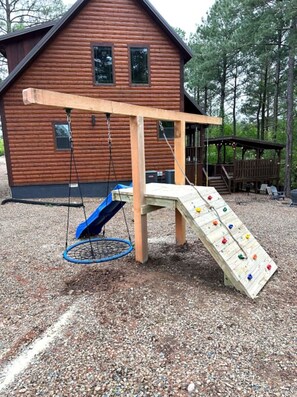 A wooden play structure with a slide and swings in front of a log cabin.