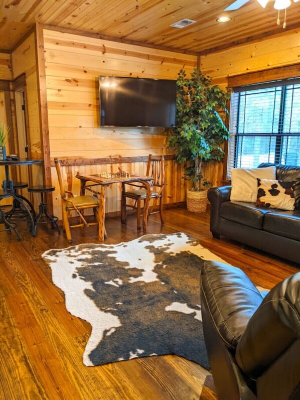 Un acogedor interior de cabaña con detalles de madera, un televisor de pantalla plana en la pared, una mesa de comedor con sillas, un sofá de cuero y una planta grande en maceta.