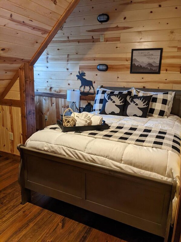 A cozy wooden cabin bedroom with a bed, checkered bedding, a moose-themed pillow, a lamp, and a framed picture on the wall.