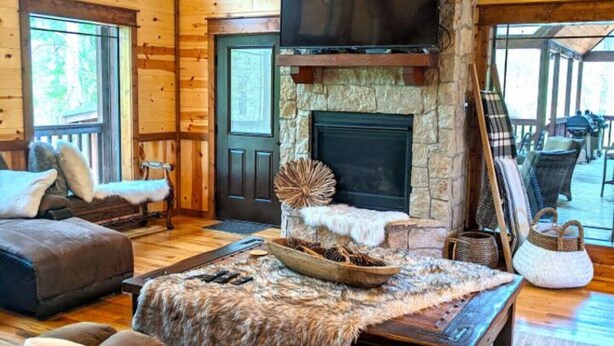 A spacious living room with a stone fireplace, wooden ceiling beams, and a large TV mounted above it.