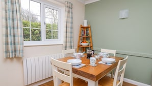 A dining area with a wooden table and white chairs, a radiator, a window with curtains, and a bookshelf with books and a fruit bowl. - Oak Tree Cottage (Cardigan)