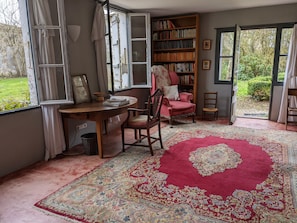 A room with a large window, a wooden desk, a red armchair, a bookshelf, and a patterned rug.
