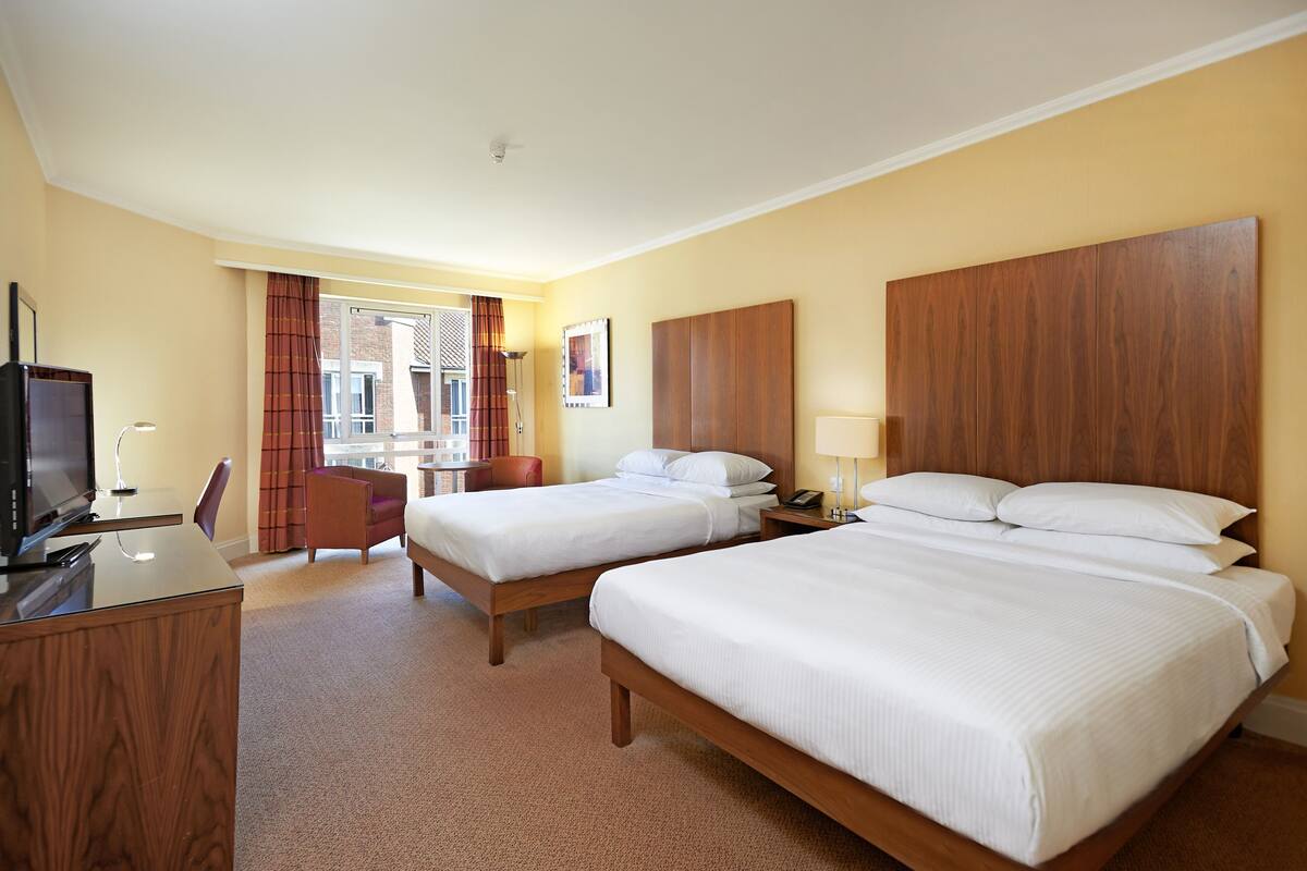Ein Hotelzimmer mit zwei Betten, einem Schreibtisch, einem Sessel, einem Fernseher und einem Fenster mit Vorhängen.