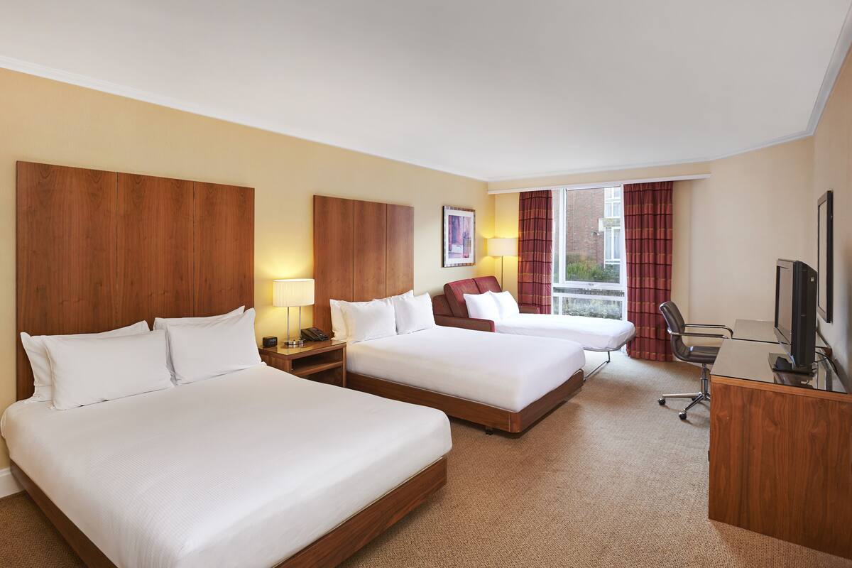 Ein Hotelzimmer mit zwei Betten, einem Schreibtisch, einem Stuhl, einem Fernseher und einem Fenster mit Vorhängen.