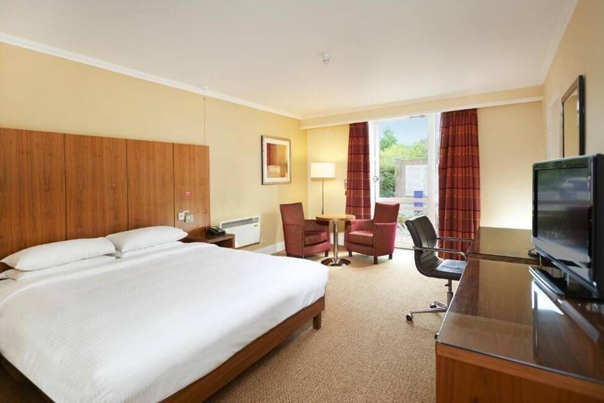 Ein Hotelzimmer mit einem großen Bett, einem Schreibtisch mit Fernseher, einem Sessel, einem Tisch und einem Fenster mit Vorhängen.