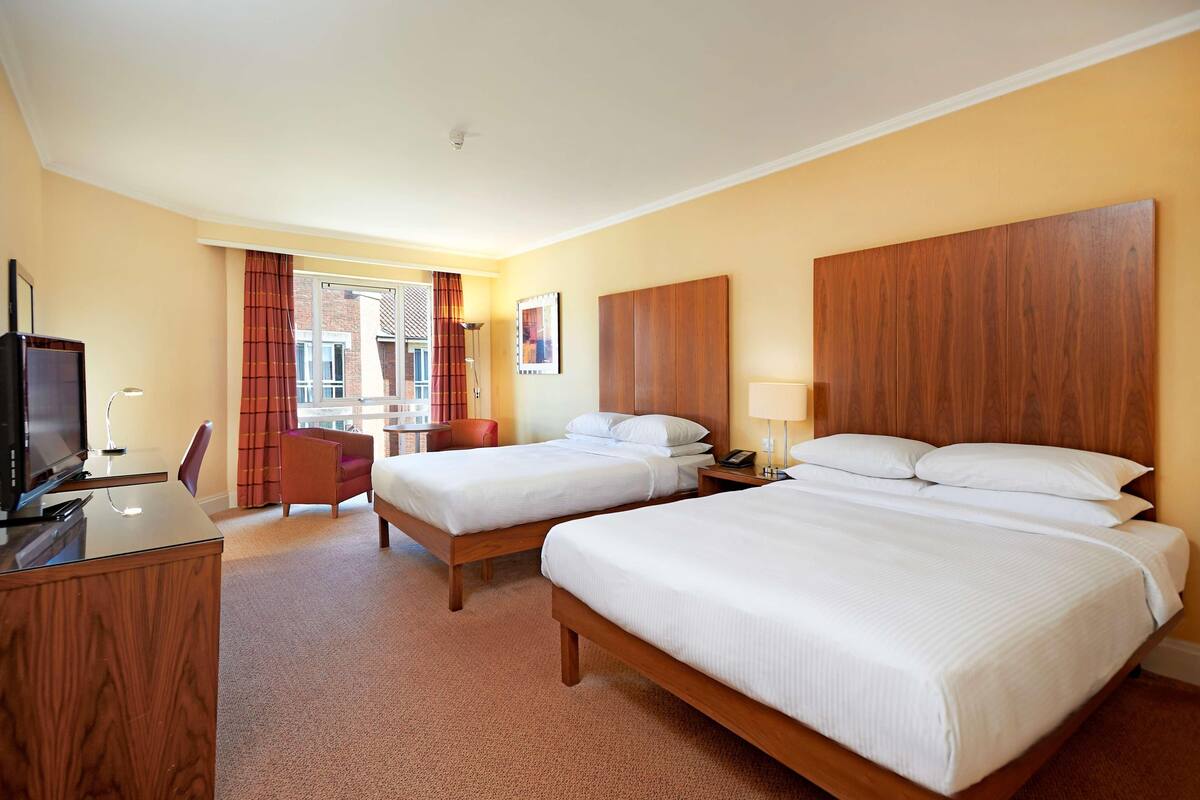 A hotel room with two beds, a desk, a chair, a TV, and a window with curtains.