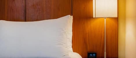 Una cama bien tendida con sábanas blancas, cabecera de madera, mesita de noche con lámpara y un reloj digital que marca las 11:19.