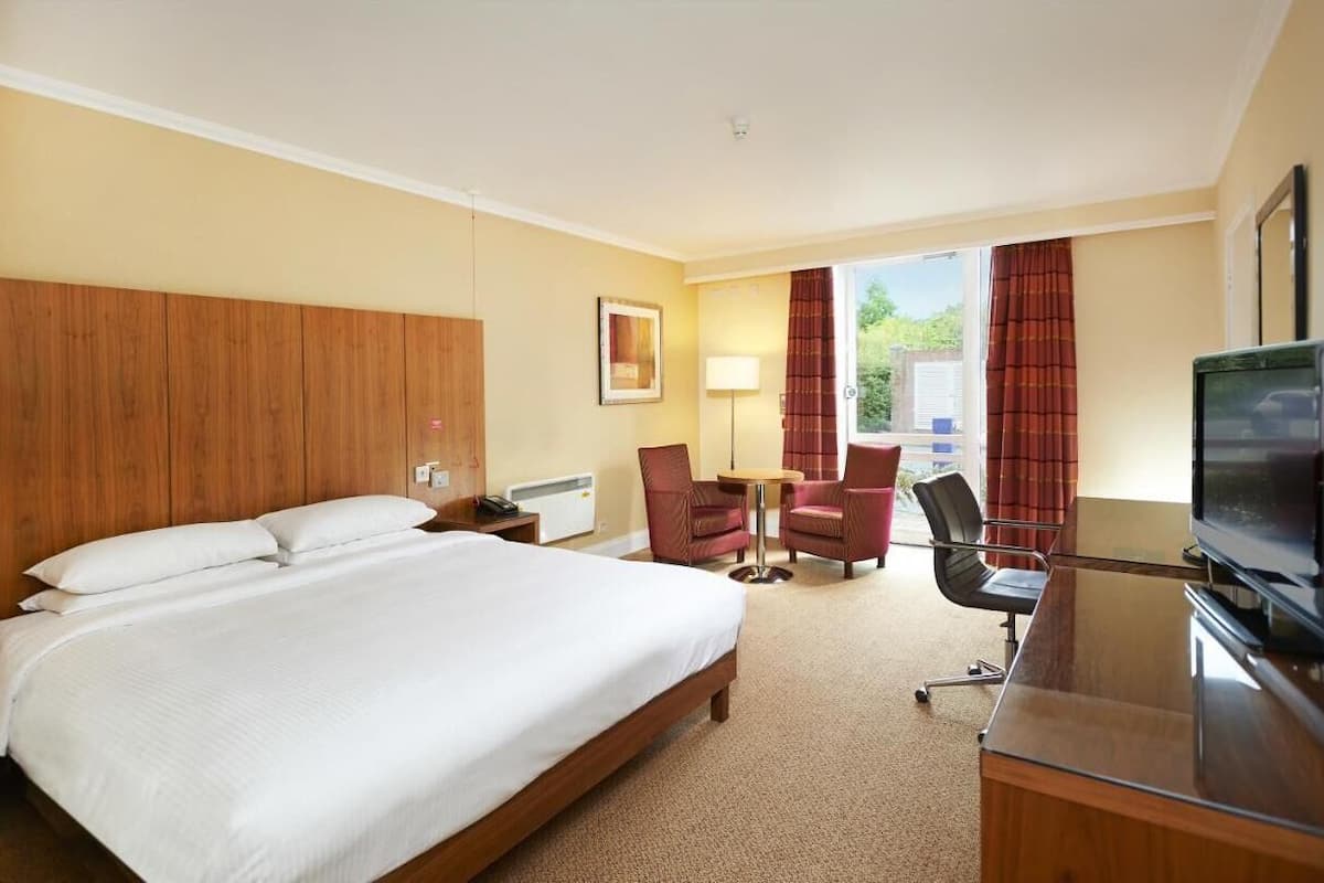 Ein Hotelzimmer mit einem großen Bett, einem Schreibtisch mit Fernseher, zwei Stühlen, einer Lampe und einem fenster mit Vorhängen.