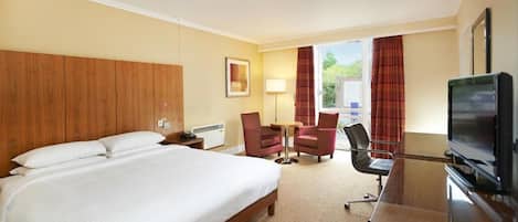 A hotel room with a large bed, a desk with a television, two chairs, a lamp, and a window with curtains.