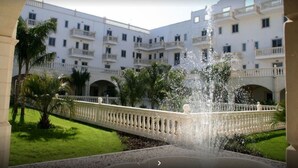 Un patio con una fuente, palmeras y un edificio blanco con balcones.