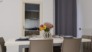 Un comedor con una mesa lista para dos personas, un florero con flores y vistas a una cocina por una puerta abierta.