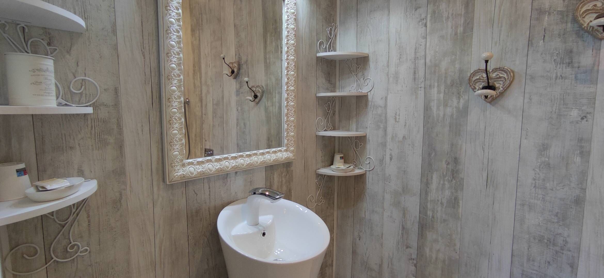 Un baño con pared de madera, un lavamanos blanco, un espejo con marco decorativo y apliques de pared ornamentales.