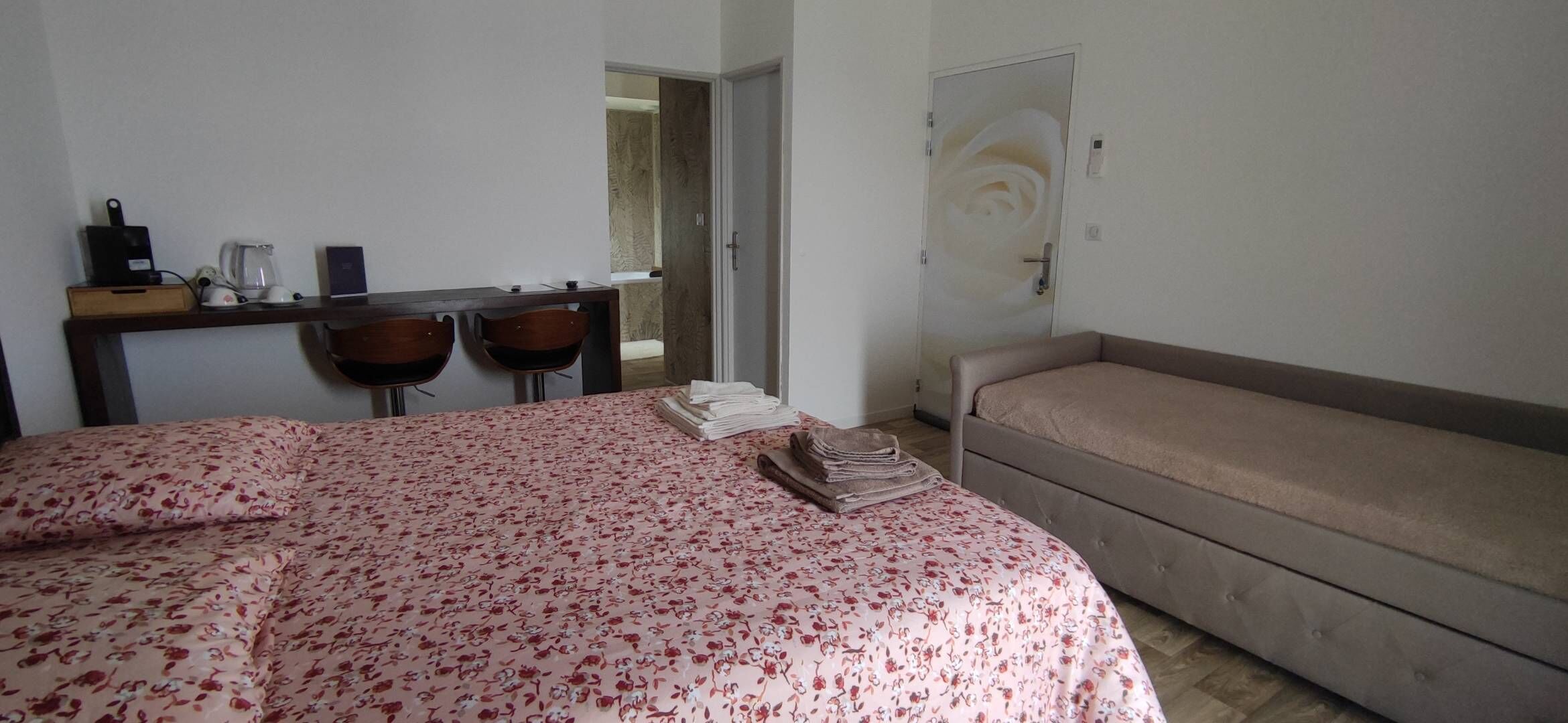 Un dormitorio con cama, sofá, escritorio con sillas, cafetera y puerta decorada con un diseño de rosa.
