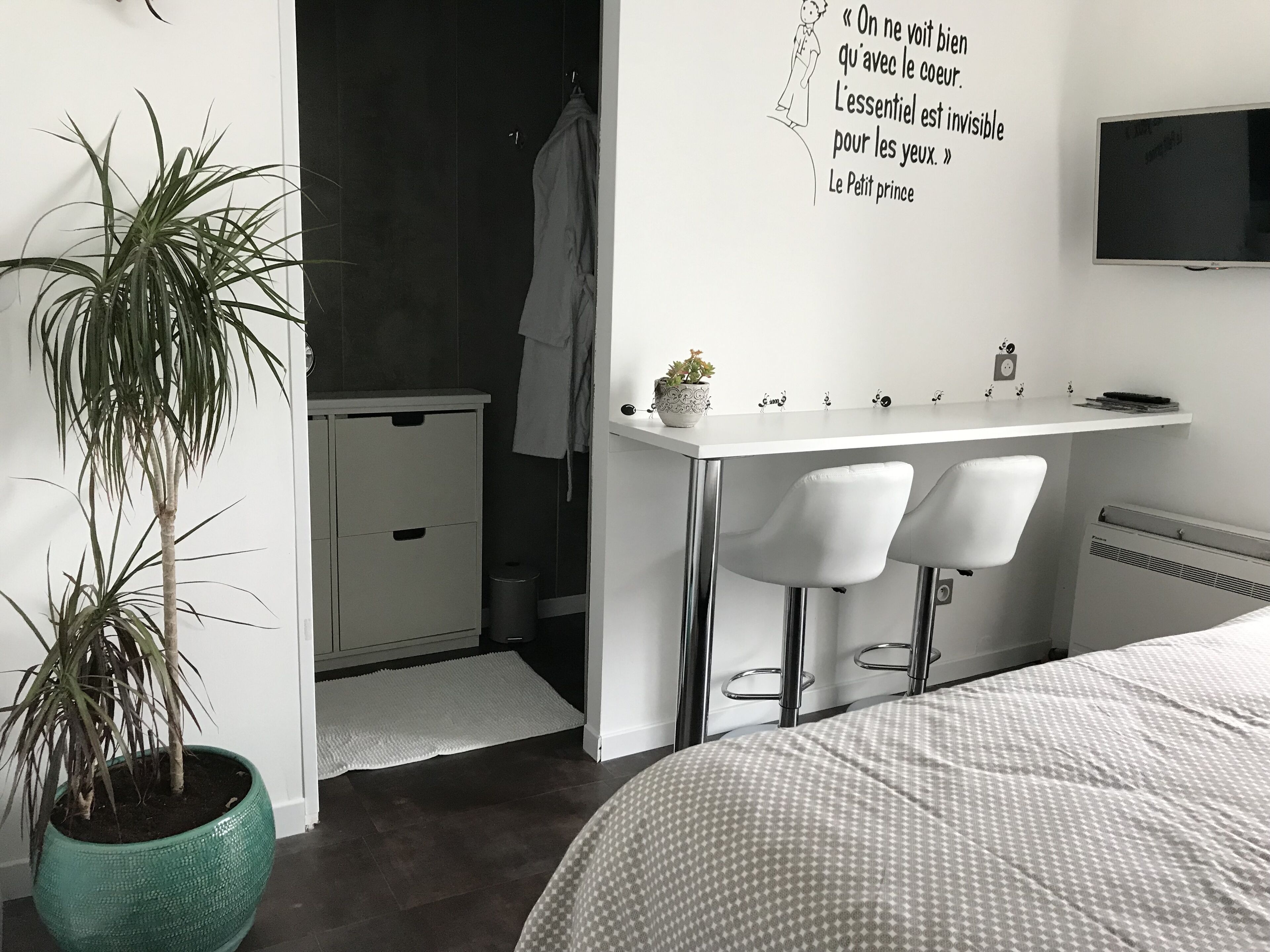 Un dormitorio con cama, escritorio con dos sillas, planta en maceta, televisor montado en la pared y una cita en la pared.