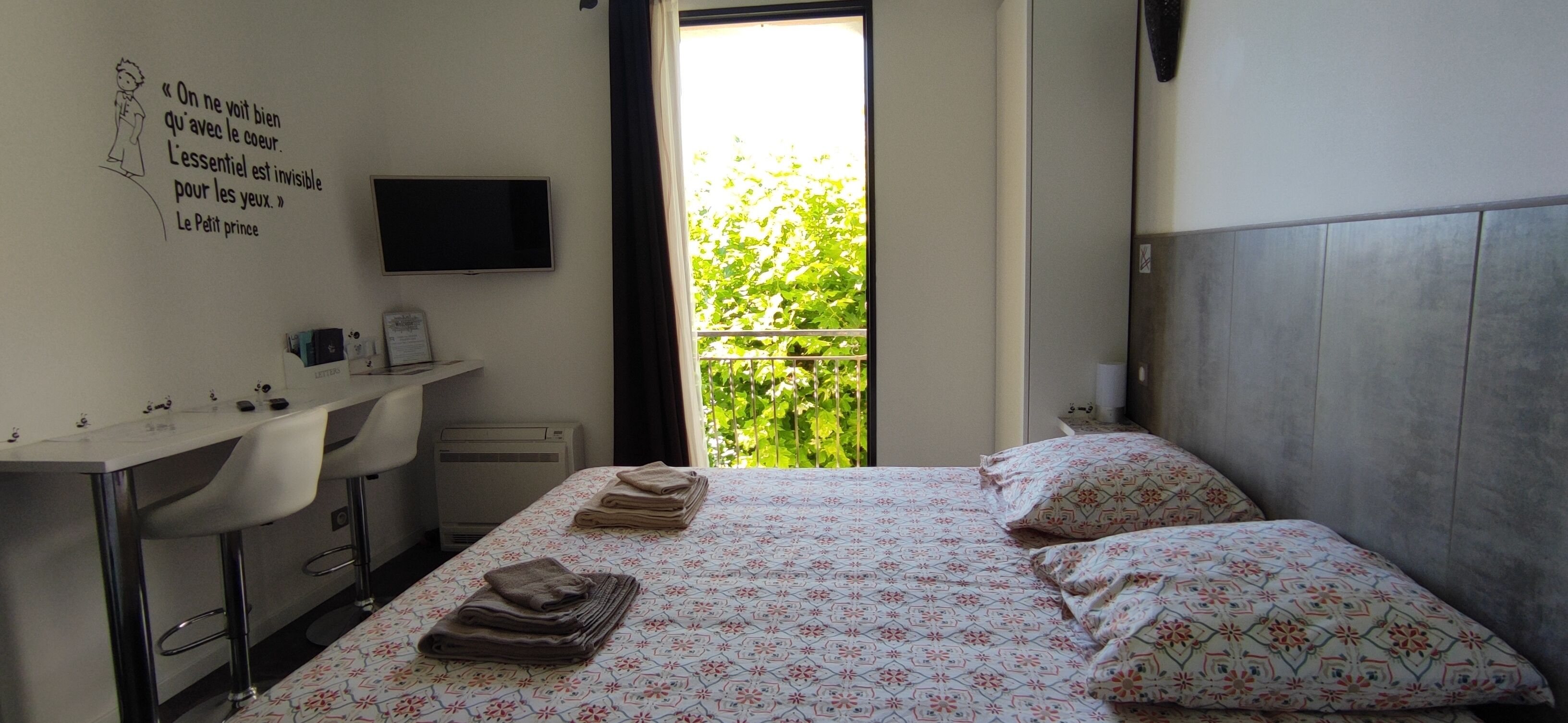 A hotel room with a bed, a desk, a chair, a TV, and a window with a view of greenery.