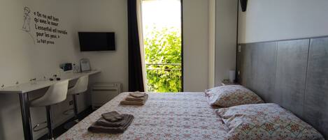 A hotel room with a bed, a desk, a chair, a TV, and a window with a view of greenery.