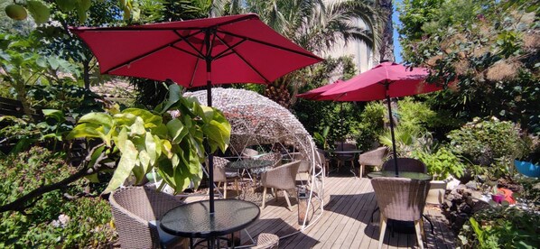 Un patio al aire libre con mobiliario de mimbre, sombrillas rojas y abundante vegetación.