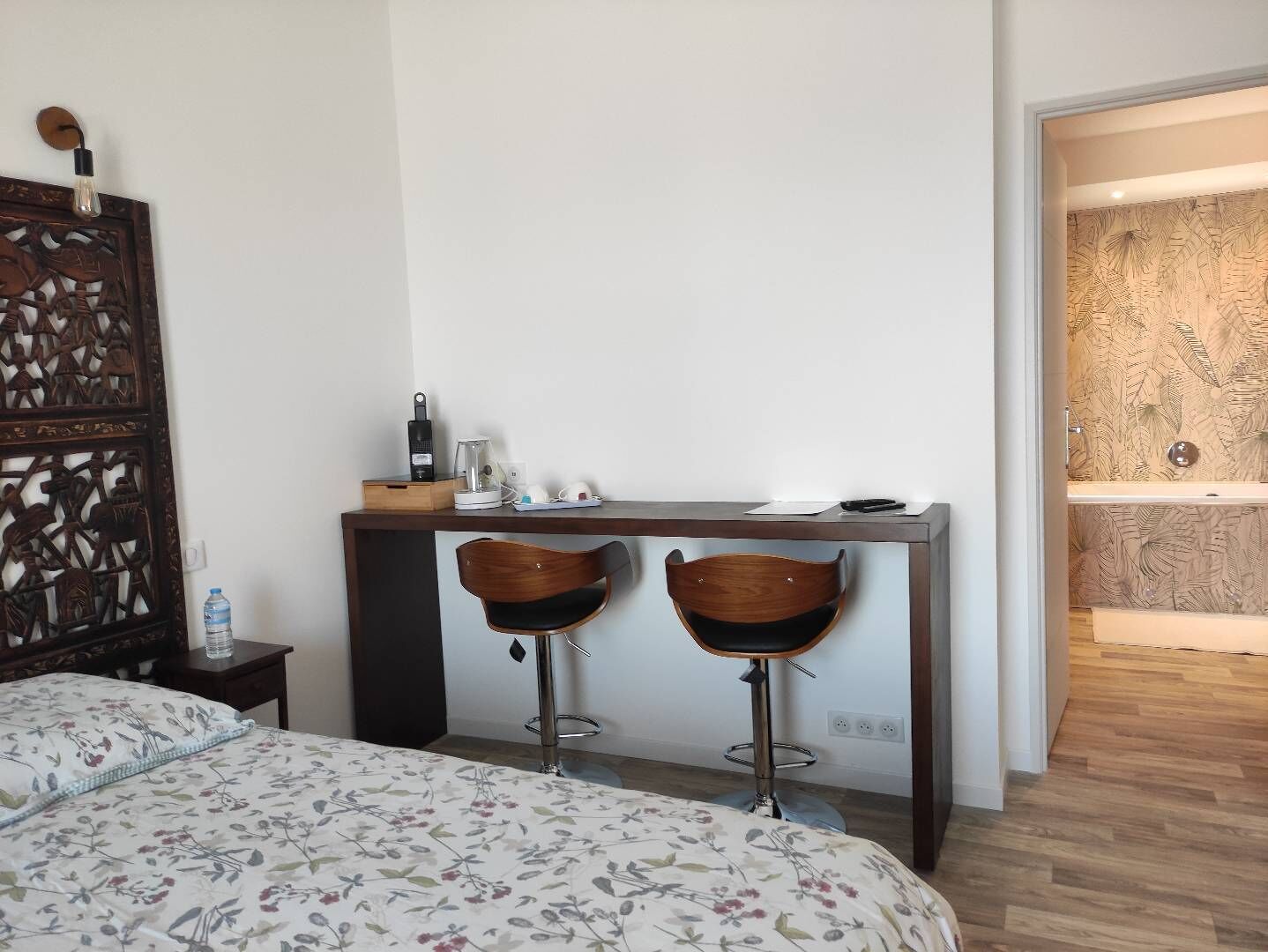 Habitación de hotel con cama, escritorio con dos taburetes altos, cafetera y un panel decorativo en la pared.