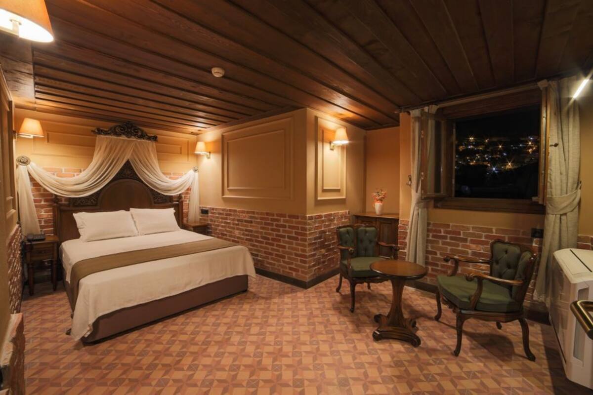 A hotel room with a large bed, a brick accent wall, a small table, two chairs, and a window with a city view at night.