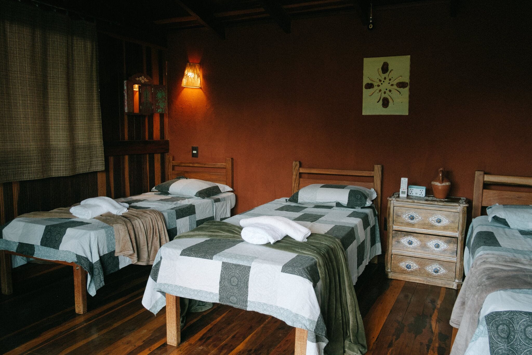 A room with three wooden beds, each with a quilted bedspread and pillows. There is a wooden nightstand with a lamp and a small decorative item on top. The room has wooden flooring and a wall with a single piece of wall art.