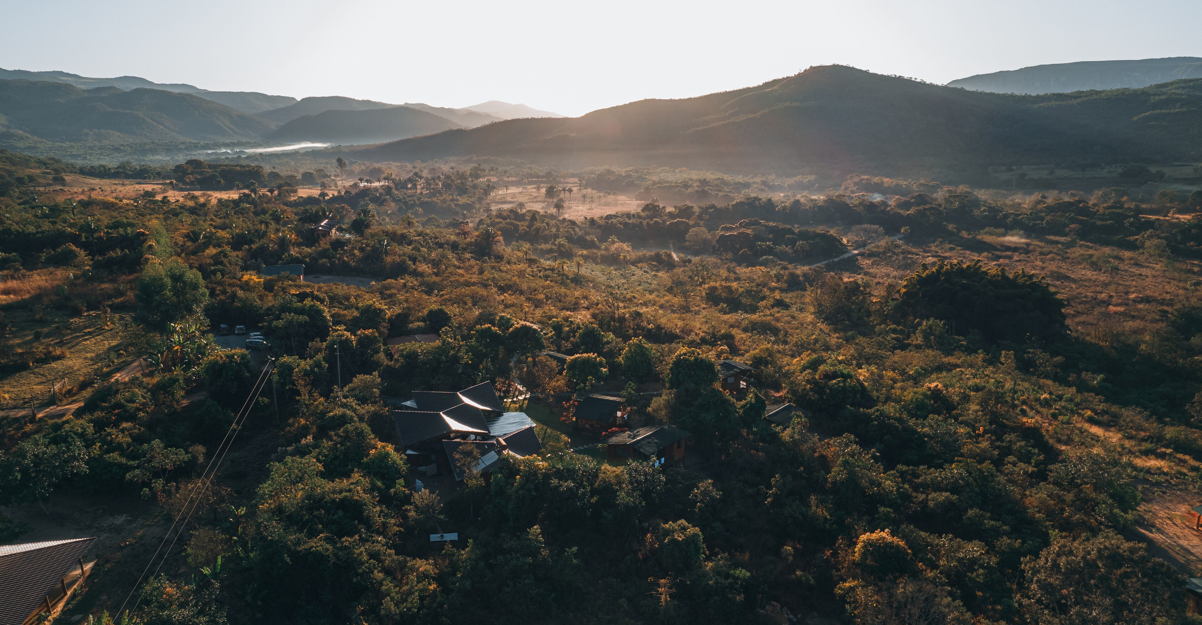Vista aérea de uma área rural com casas dispersas, cercada por vegetação densa e colinas ao fundo.