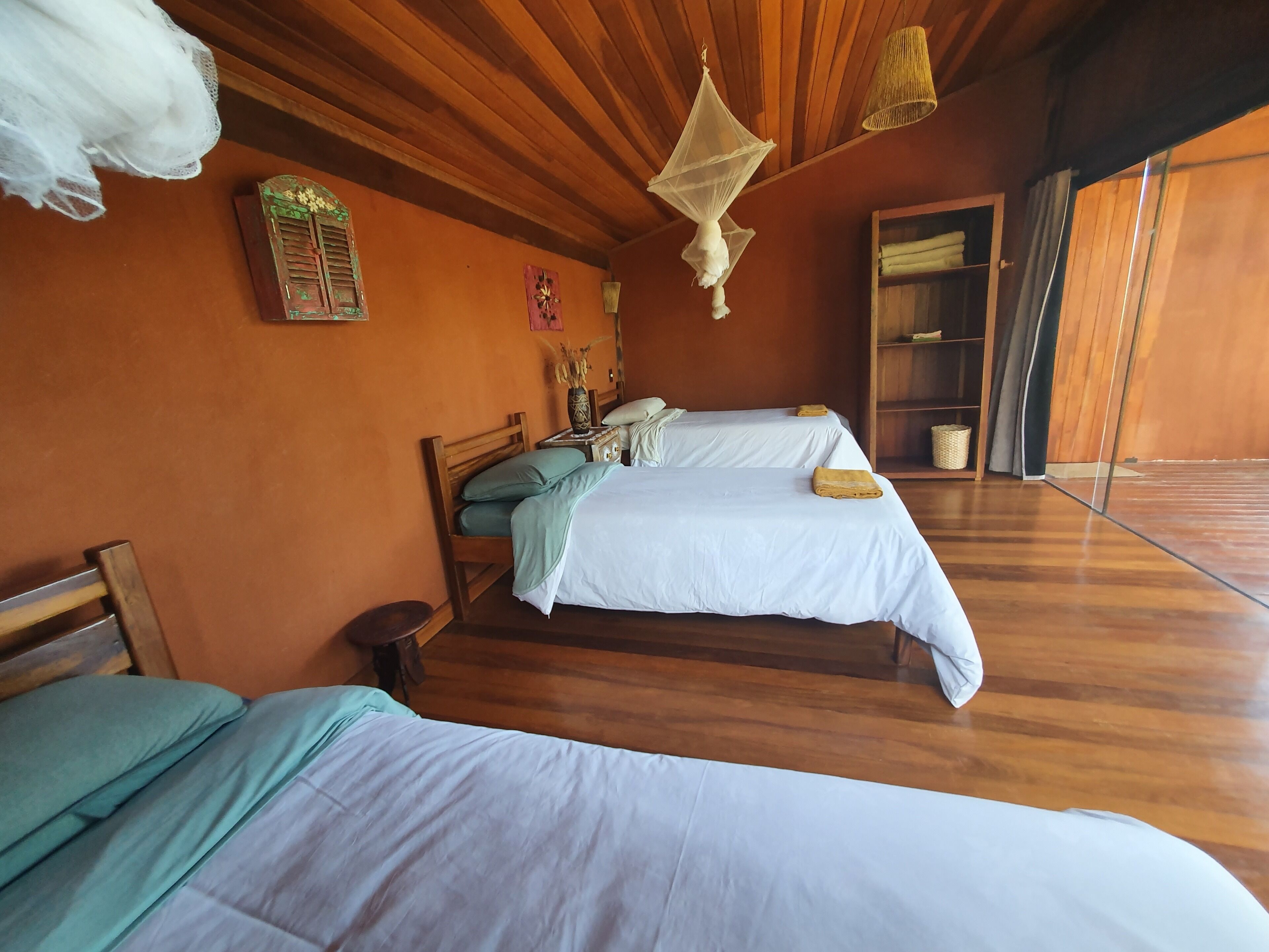Una habitación con dos camas, suelo de madera, techo de madera y una lámpara de techo tejida, además de una puerta de madera.