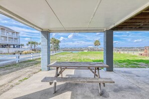 A covered outdoor area with a picnic table, a view of a beach, and a building in the background.