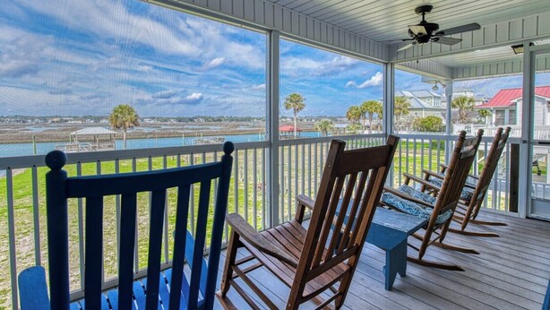 A covered porch with wooden chairs and a table, overlooking a body of water and distant buildings. - Inlet Joy 2nd Row Channel Front (Garden City Beach)