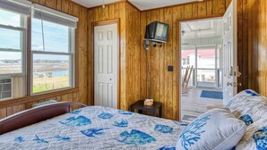 A bedroom with wooden paneling, a bed with a blue and white quilt, a television on the wall, and a view of a deck and outdoor area. - Inlet Joy 2nd Row Channel Front (Garden City Beach)