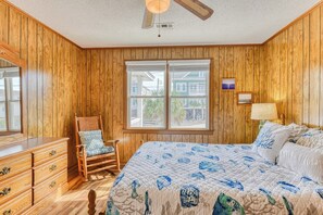A bedroom with wooden paneling, a bed with a blue floral quilt, a rocking chair, a dresser, and a window with a view of houses.