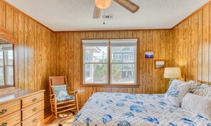 A bedroom with wooden paneling, a bed with a floral quilt, a rocking chair, a dresser, a window with a view of houses, and a ceiling fan.